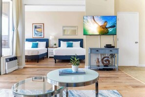 Quarto de hotel com cama, televisão, mesa de cabeceira, uma planta pequena e uma mesinha de centro de vidro.