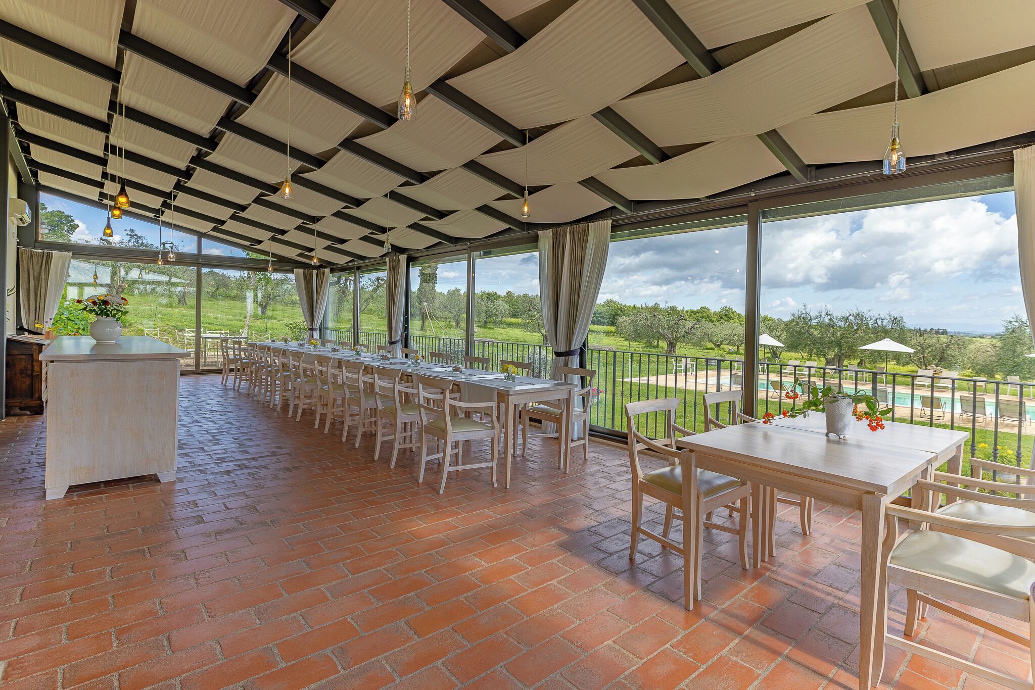 Un espace extérieur abrité, aménagé pour les repas, avec un sol en briques, des tables, des chaises et des lampes suspendues.