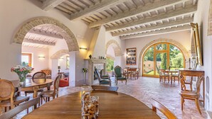 A spacious dining area with wooden tables and chairs, a fireplace, and large windows offering a view of the outdoors. - LE BUCHE ESTATE  VILLA RENTAL  (Sarteano)