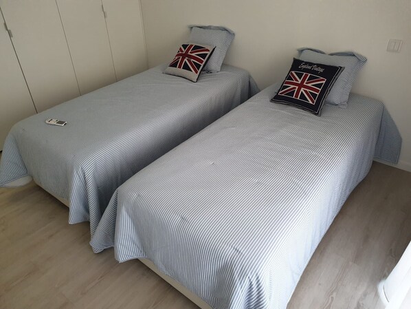 Deux lits simples avec une literie à rayures et des oreillers ornés d’un motif Union Jack, dans une pièce dotée d’un plancher en bois et de murs blancs.