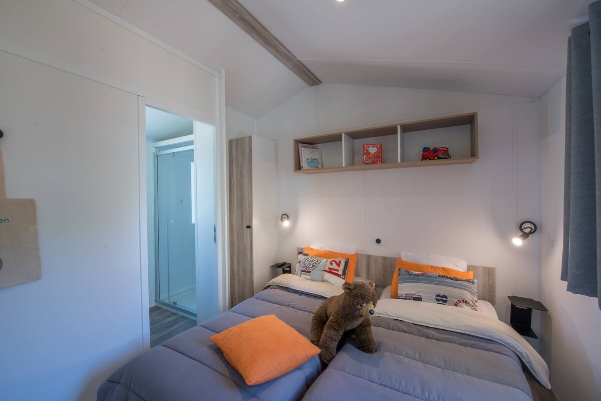 Un dormitorio con cama, almohadas, un osito de peluche, una mesita de noche, un estante con libros y juguetes, y una puerta a un pasillo.