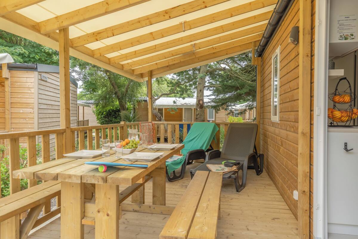 Una terraza de madera con mesas y sillas de picnic, un espacio cubierto con pérgola y un edificio pequeño al fondo.