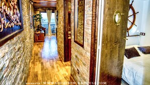 A hallway with wooden flooring and stone walls, leading to a bedroom with a bed and a ship's wheel on the wall. - Seafront rental by Thierry Bourgait (Banyuls-sur-Mer)