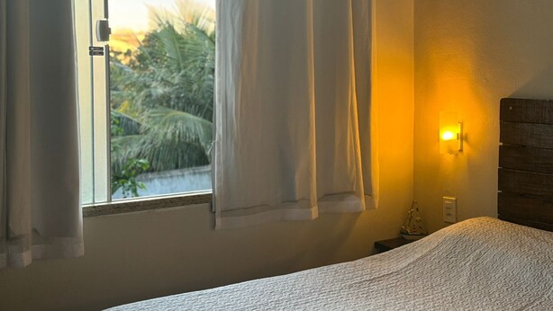 A bedroom with a bed, a window with curtains, a wall-mounted light, and a view of palm trees outside. - Suíte em Geribá, Próximo a Praia (Armação do Búzios)