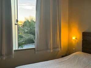 Una camera da letto con un letto, una finestra con tende, una lampada a parete e vista su palme.