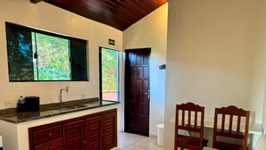 A kitchen with a large window, a sink, and wooden cabinets. There are two wooden chairs and a white table in the living area. - Suíte em Geribá, Próximo a Praia (Armação do Búzios)