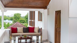 Eine überdachte Veranda mit Holzboden, ein beiges Sofa mit roten und schwarzen Kissen, ein kleiner Tisch mit einer Pflanze sowie der Blick auf ein Gebäude mit rotem Dach und viel Grün.
