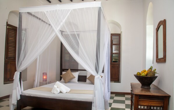 A bed with a canopy, pillows, a bedside table with a fruit bowl, and a mirror on the wall. - The Manor House by Sansi (Zanzibar Town)