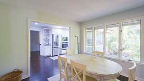 A dining area with a round table and chairs, a kitchen in the background, and large windows with a view of greenery.