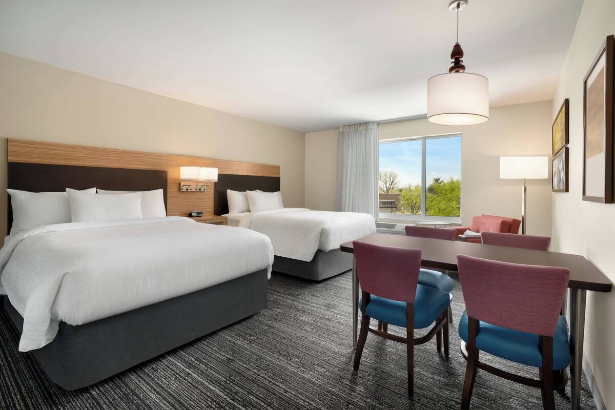 A hotel room with two beds, a desk, chairs, and a large window with a view.