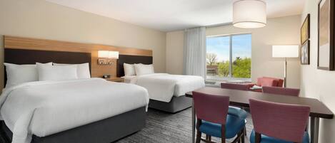 A hotel room with two beds, a desk, chairs, and a large window with a view.