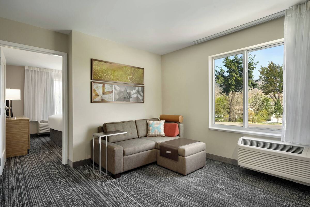 A hotel room with a sectional sofa, a small table, a window with a view of trees, and a radiator.