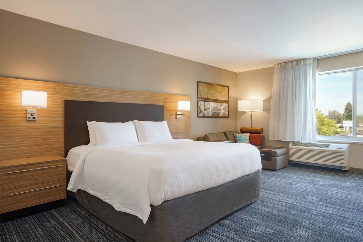 A hotel room with a large bed, bedside tables, a lamp, a window with curtains, and a wall-mounted air conditioner.