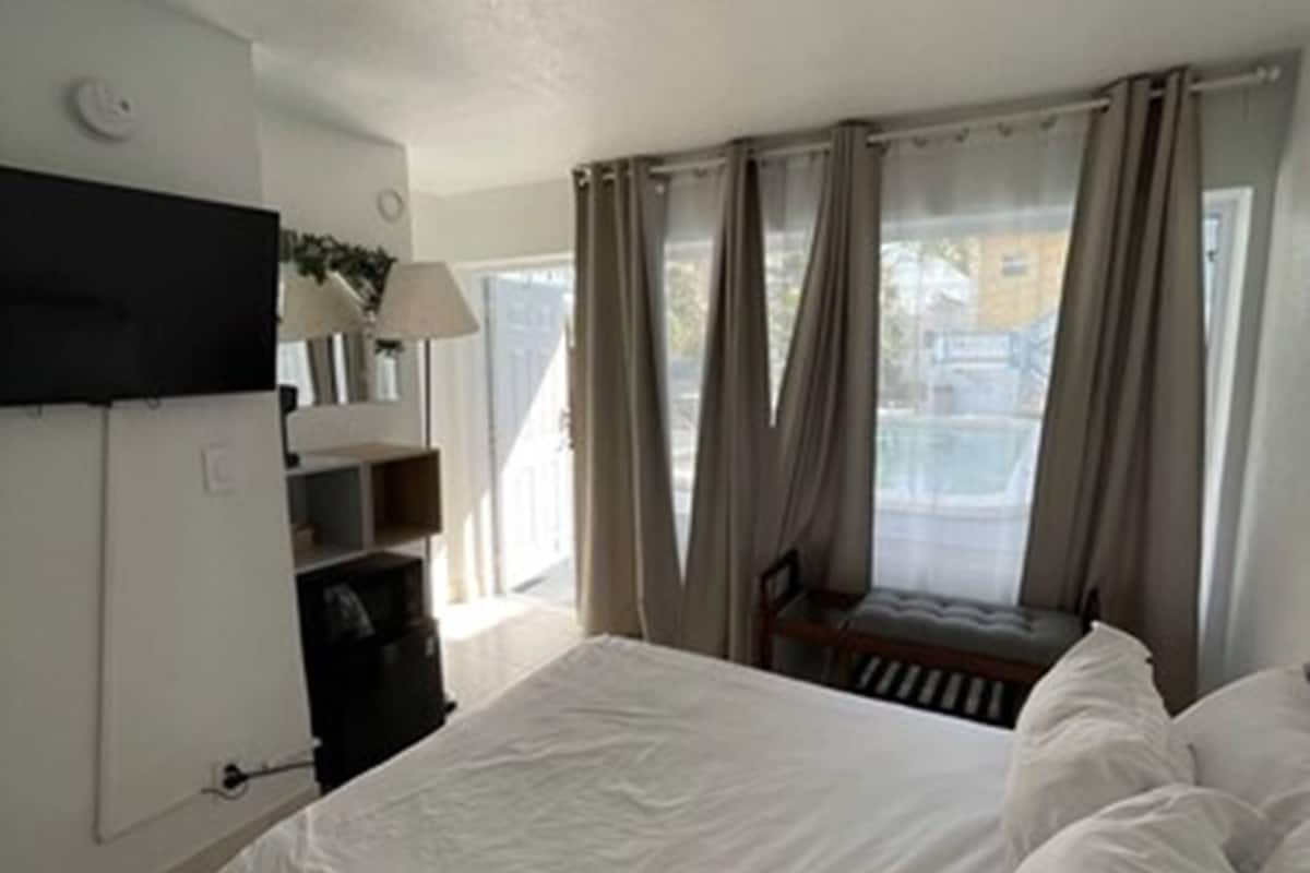 A bedroom with a bed, a TV mounted on the wall, a window with curtains, and a view of the outside.