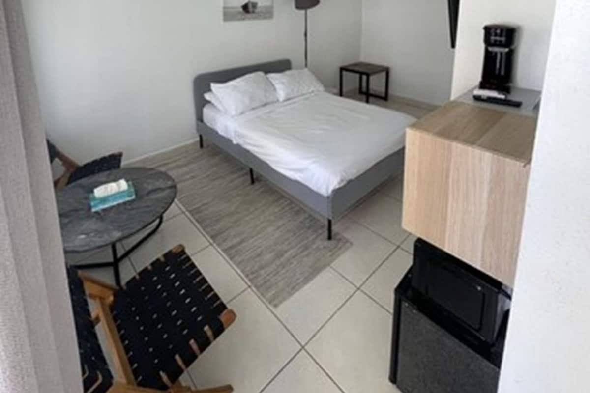 A small, modern bedroom with a bed, a desk, a chair, a lamp, and a coffee machine.