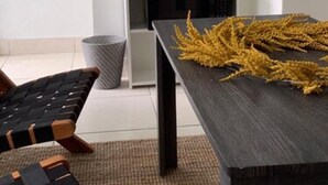 Ein modernes Wohnzimmer mit einem dunklen Holztisch, einer gewebten Bank, einer Topfpflanze und einer Kaffeemaschine auf einem weißen Regal.