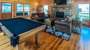 A wooden cabin interior with a pool table, leather sofas, a stone fireplace, and large windows.