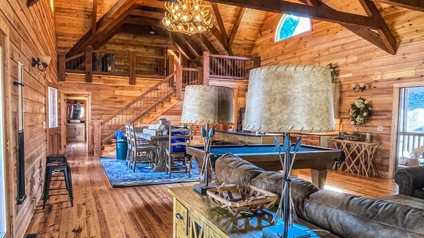 A spacious log cabin interior with a high ceiling, wooden beams, a central staircase, a dining area with a table and chairs, and a large window providing natural light.