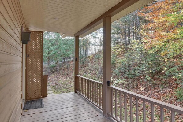 Une terrasse en bois avec une balustrade, une porte ornée d’un motif en treillis et une vue sur une zone boisée aux couleurs automnales.