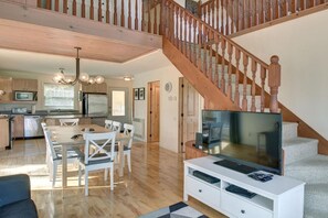 Un vaste espace de vie agrémenté d’un escalier en bois, d’une table à manger avec des chaises, d’une cuisine équipée d’appareils modernes et d’un téléviseur à écran plat posé sur un meuble de télévision blanc.