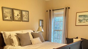Ein Schlafzimmer mit einem großen Bett, einem Nachttisch, einer Lampe und einem Fenster mit Vorhängen. An der Wand hängen gerahmte Bilder.