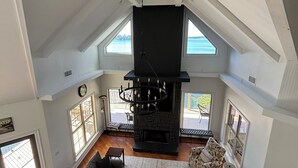 Ein geräumiger Loft mit hohen Decken, einem Kamin, einer Couch, einem gemusterten Sessel und großen Fenstern mit Blick ins Freie.