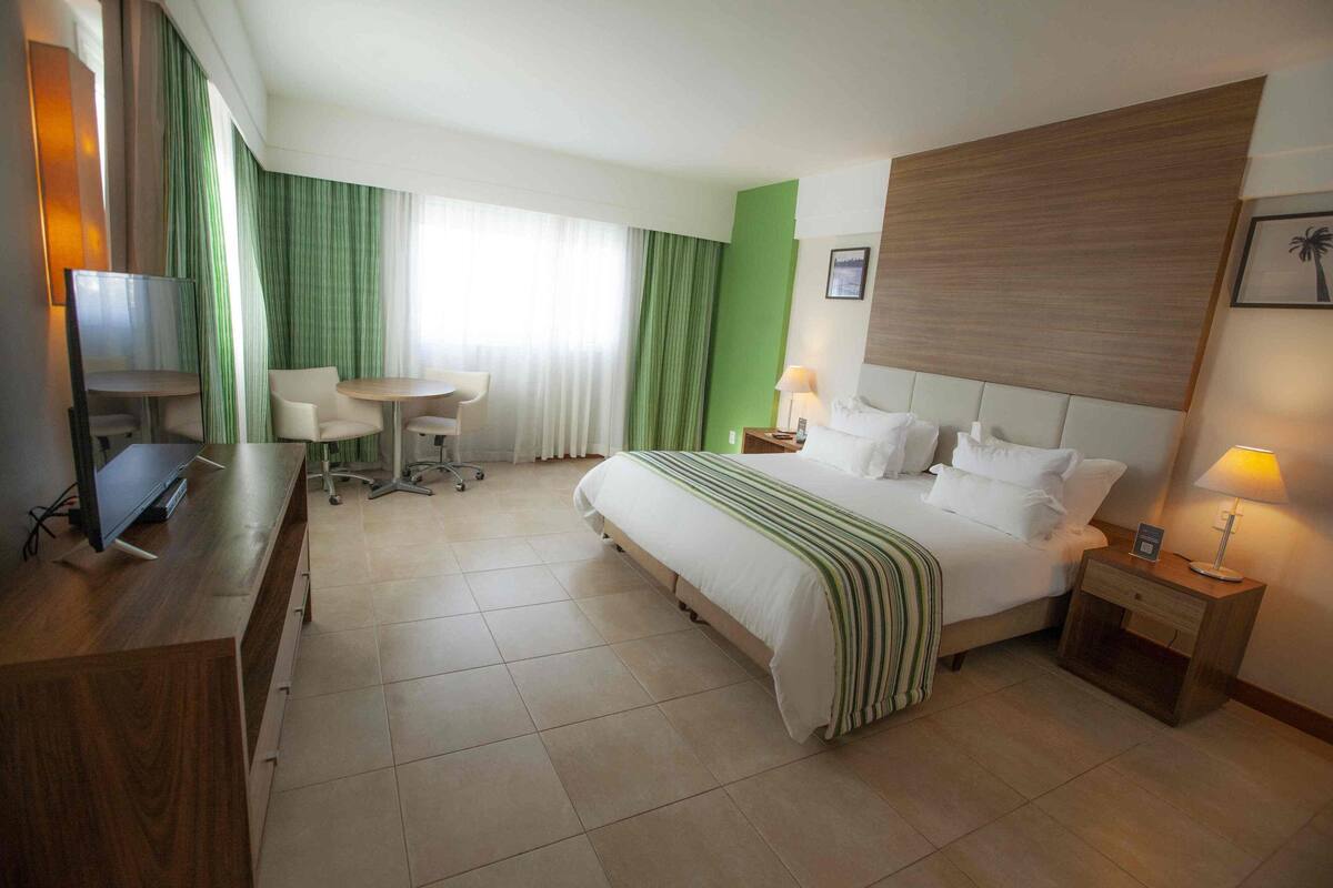 A hotel room with a bed, bedside table, lamp, desk with chair, TV, and curtains.