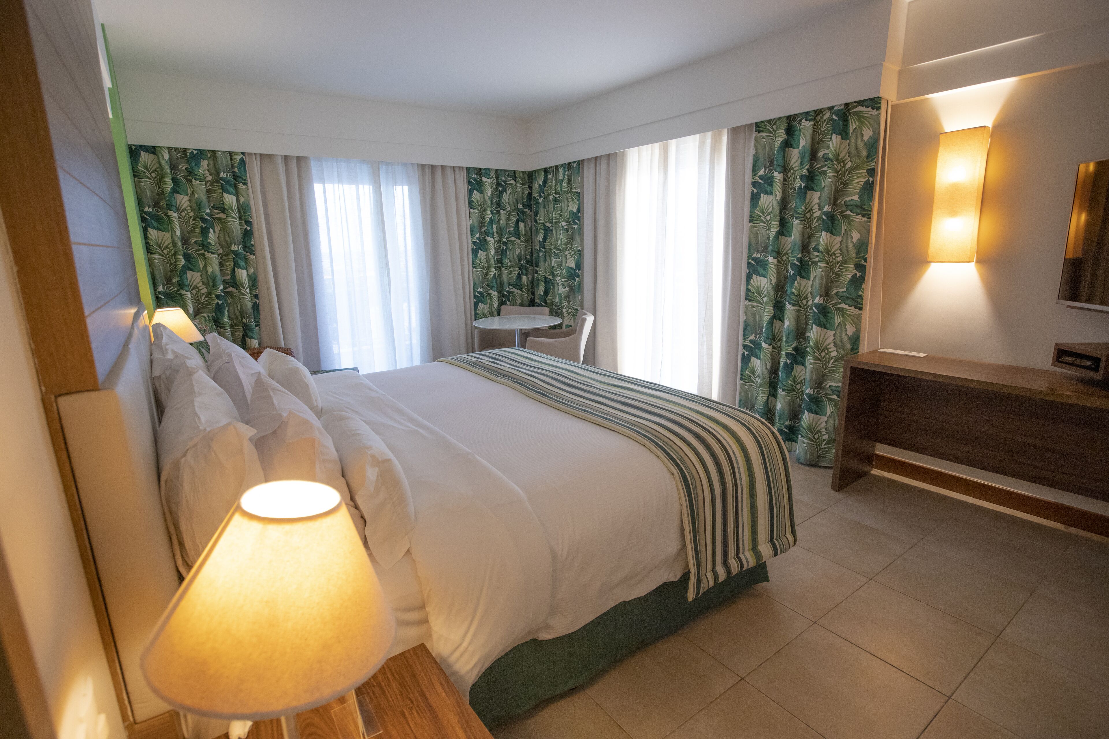 A hotel room with a large bed, a bedside lamp, a small table with a chair, and a TV.