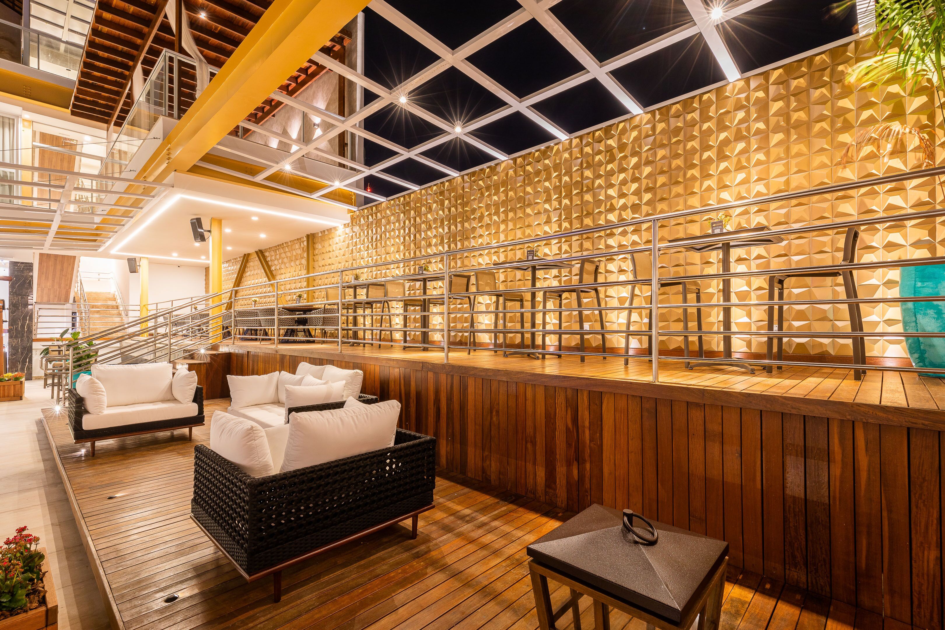 A modern indoor area with wooden flooring, white sofas, a black wicker chair, and a bar area with a wooden wall and metal railings.