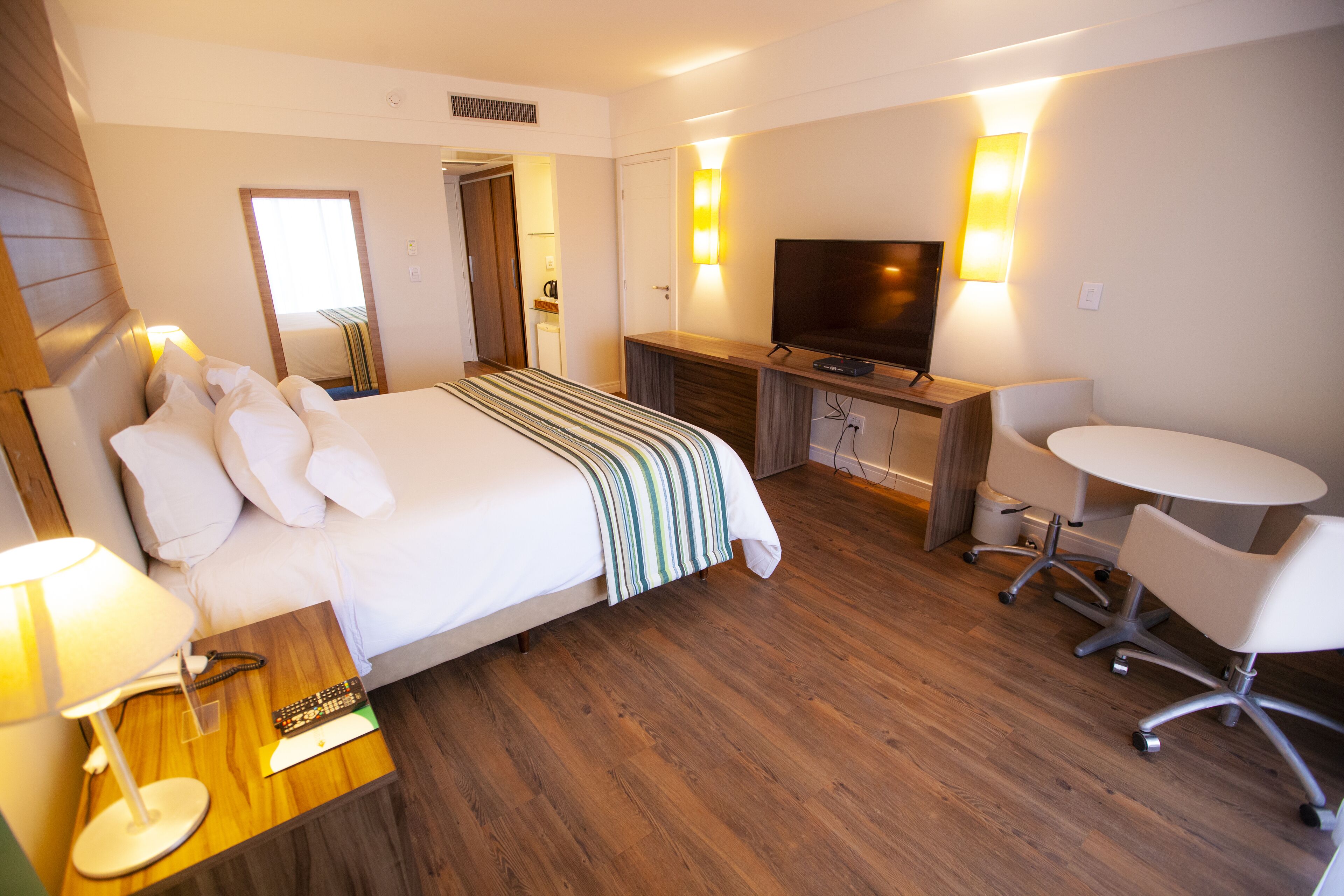 A hotel room with a bed, bedside table, lamp, TV, desk, chair, and wooden flooring.