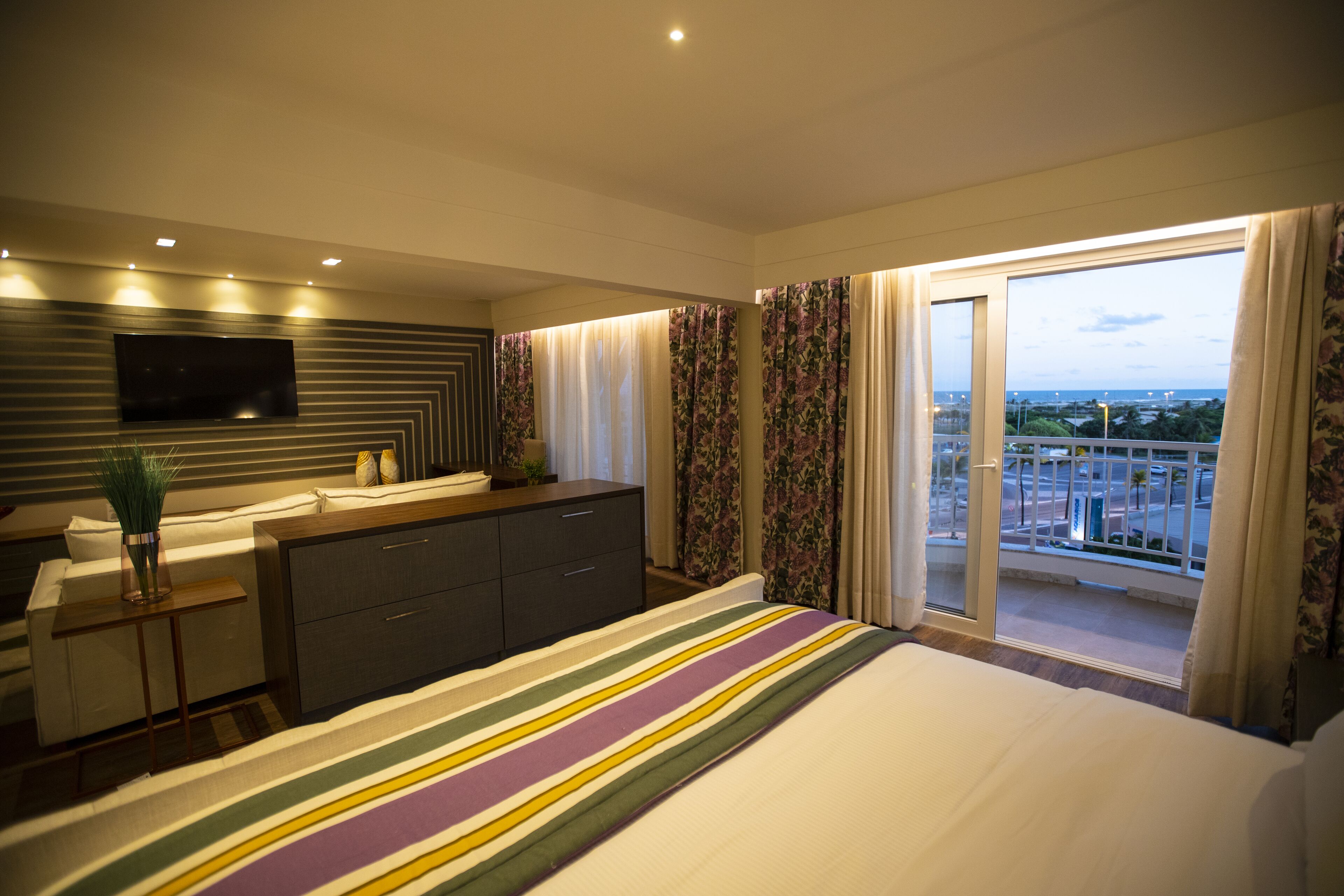 A modern hotel room with a large bed, a flat-screen TV mounted on the wall, a small bedside table with a vase, and a balcony with a city view.