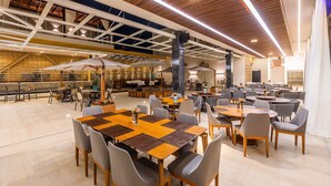 Un espacio interior amplio con varias mesas y sillas de comedor, vigas de techo de madera y un techo alto con elementos estructurales a la vista.