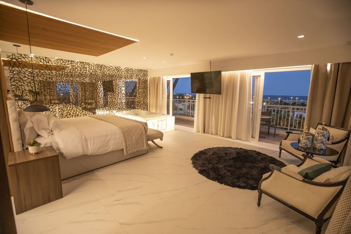 A modern bedroom with a large bed, a sitting area with a sofa and armchairs, a small table, and a balcony with a TV.