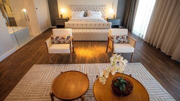 A modern hotel room with a large bed, two white armchairs, a round wooden table, and a decorative plant.