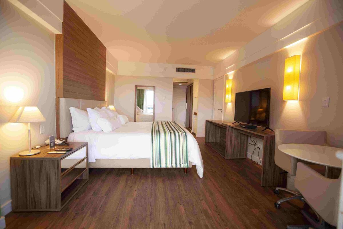 A hotel room with a bed, bedside table, TV, desk, chair, and a mirror.