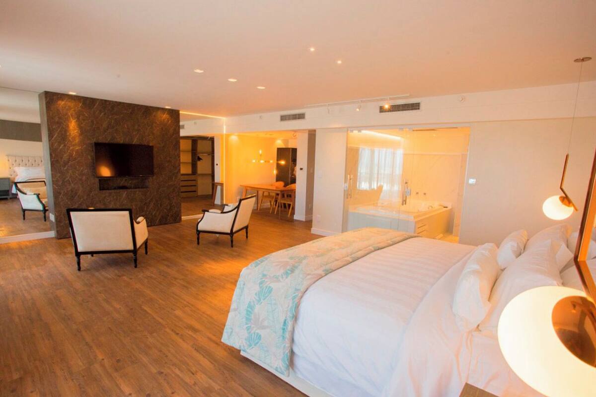 A modern hotel room with a large bed, a seating area with chairs, a TV mounted on a marble wall, and a bathroom visible through an open door.