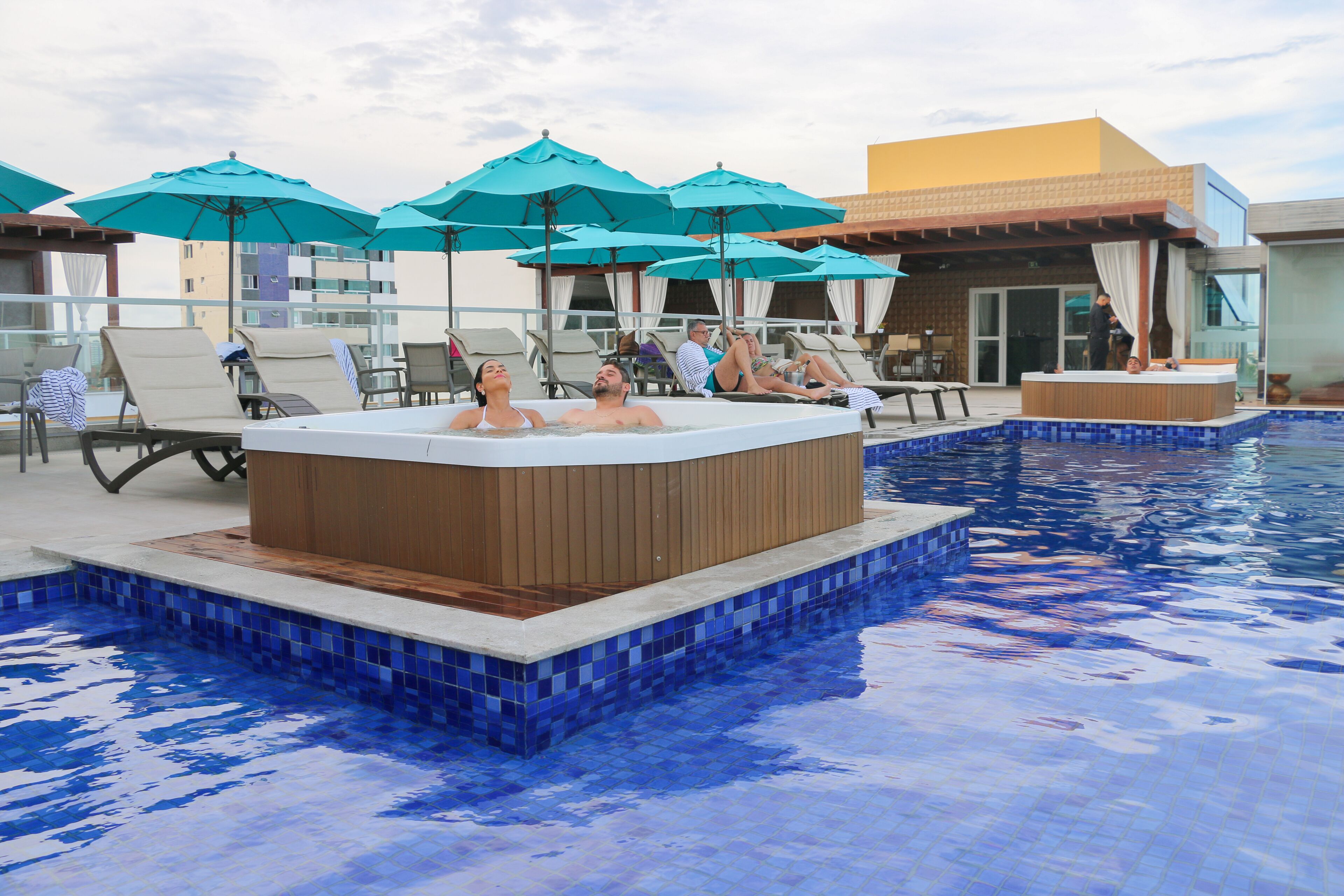 A pool area with lounge chairs and umbrellas, a hot tub, and a building with a yellow facade.