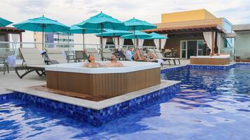 A pool area with lounge chairs and umbrellas, a hot tub, and a building with a yellow facade.