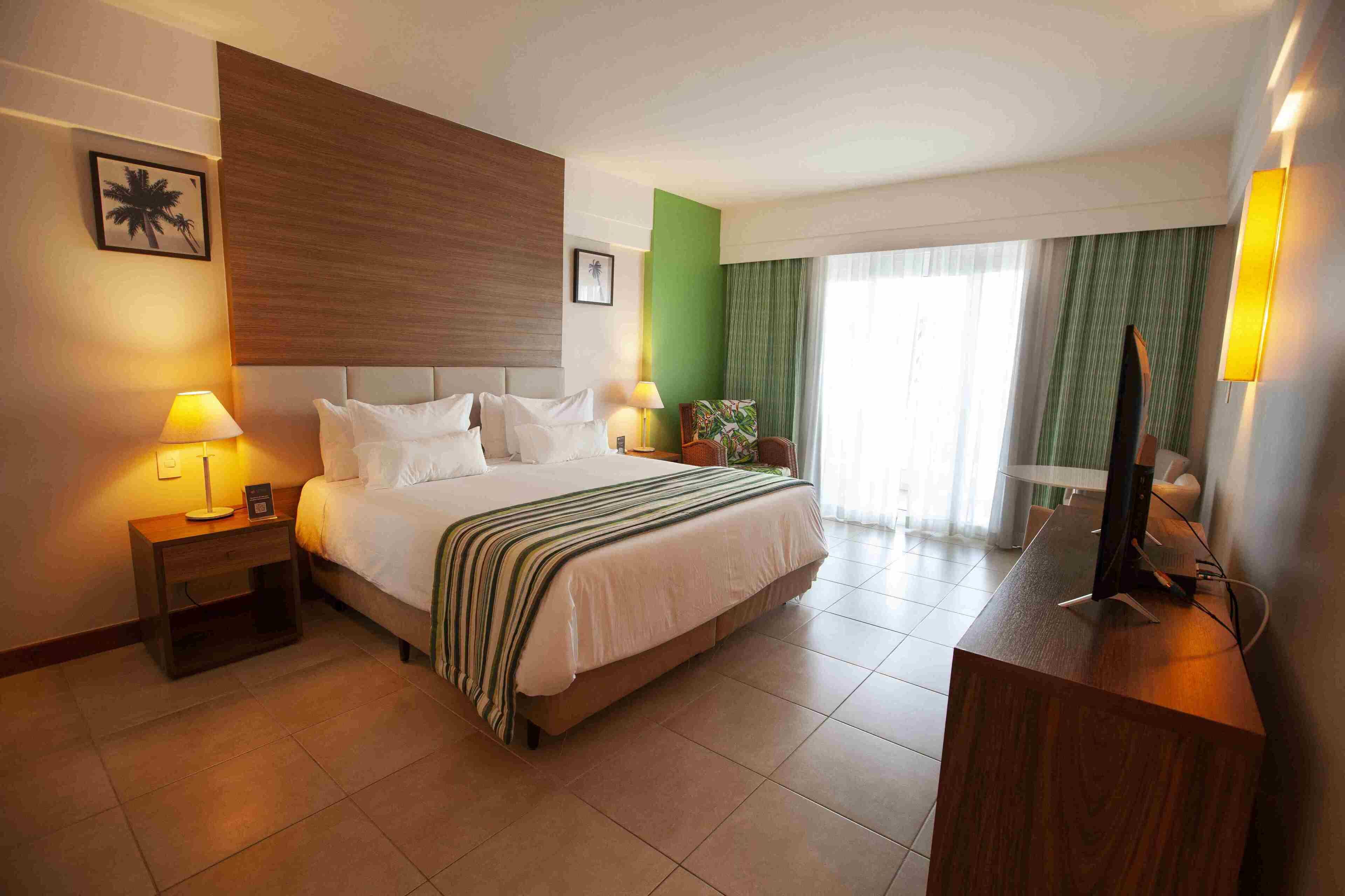 A hotel room with a bed, bedside tables, a desk, a chair, a TV, and a window with curtains.