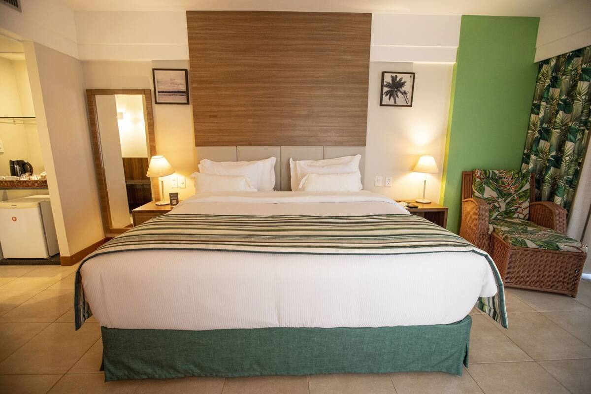A hotel room with a large bed, two bedside lamps, a small desk with a lamp, a chair, and a view of the sea.