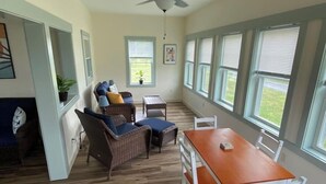 Ein Zimmer mit Esstisch, Stühlen, Korbmöbeln, einem Deckenventilator und großen Fenstern.