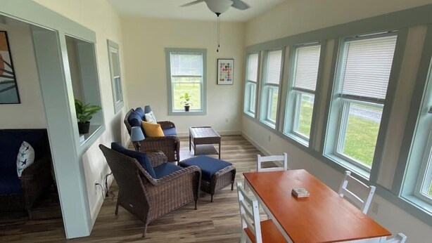 A room with a dining table, chairs, wicker furniture, a ceiling fan, and large windows. - #21 Isle View - Fishing, Sunsets, Beaches, Golfing (Alburgh)