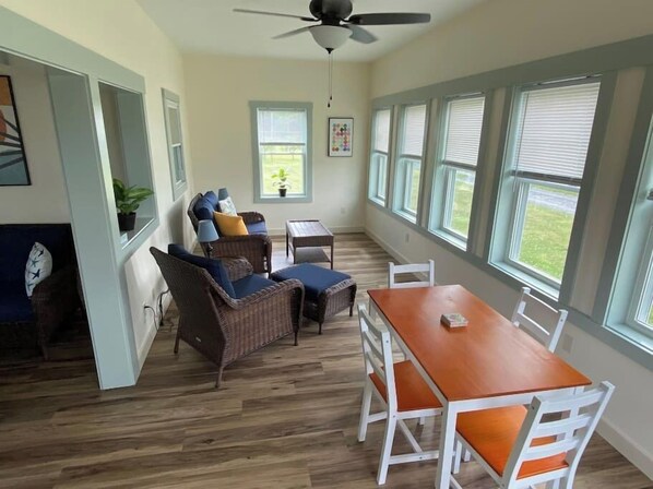 A room with a dining table, chairs, wicker furniture, a ceiling fan, and large windows.