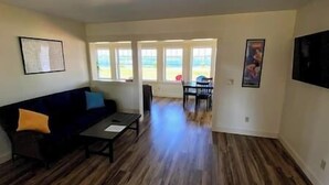 A living room with a dark-colored sofa, a small table, and a view of the outdoors through large windows. - #21 Isle View - Fishing, Sunsets, Beaches, Golfing (Alburgh)