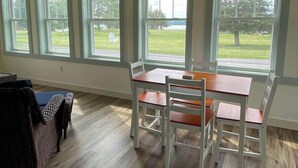 Ein Zimmer mit Esstisch, Stühlen, einem Korbstuhl und großen Fenstern mit Blick auf eine Wiese.