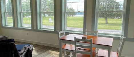 A room with a dining table, chairs, a wicker chair, and large windows with a view of a grassy area.
