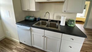 A kitchen with white cabinets, a black countertop, a stainless steel sink, and a dish rack. - #21 Isle View - Fishing, Sunsets, Beaches, Golfing (Alburgh)