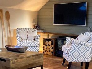 A living room with patterned armchairs, a wooden coffee table, a TV mounted on the wall, and a stack of firewood.