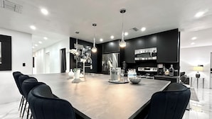 Une cuisine moderne dotée d’un grand îlot central, de chaises noires, d’appareils électroménagers en acier inoxydable et de luminaires suspendus.