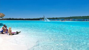 Une mer turquoise aux eaux limpides, avec un toboggan aquatique, une plage animée et une zone boisée en arrière-plan.